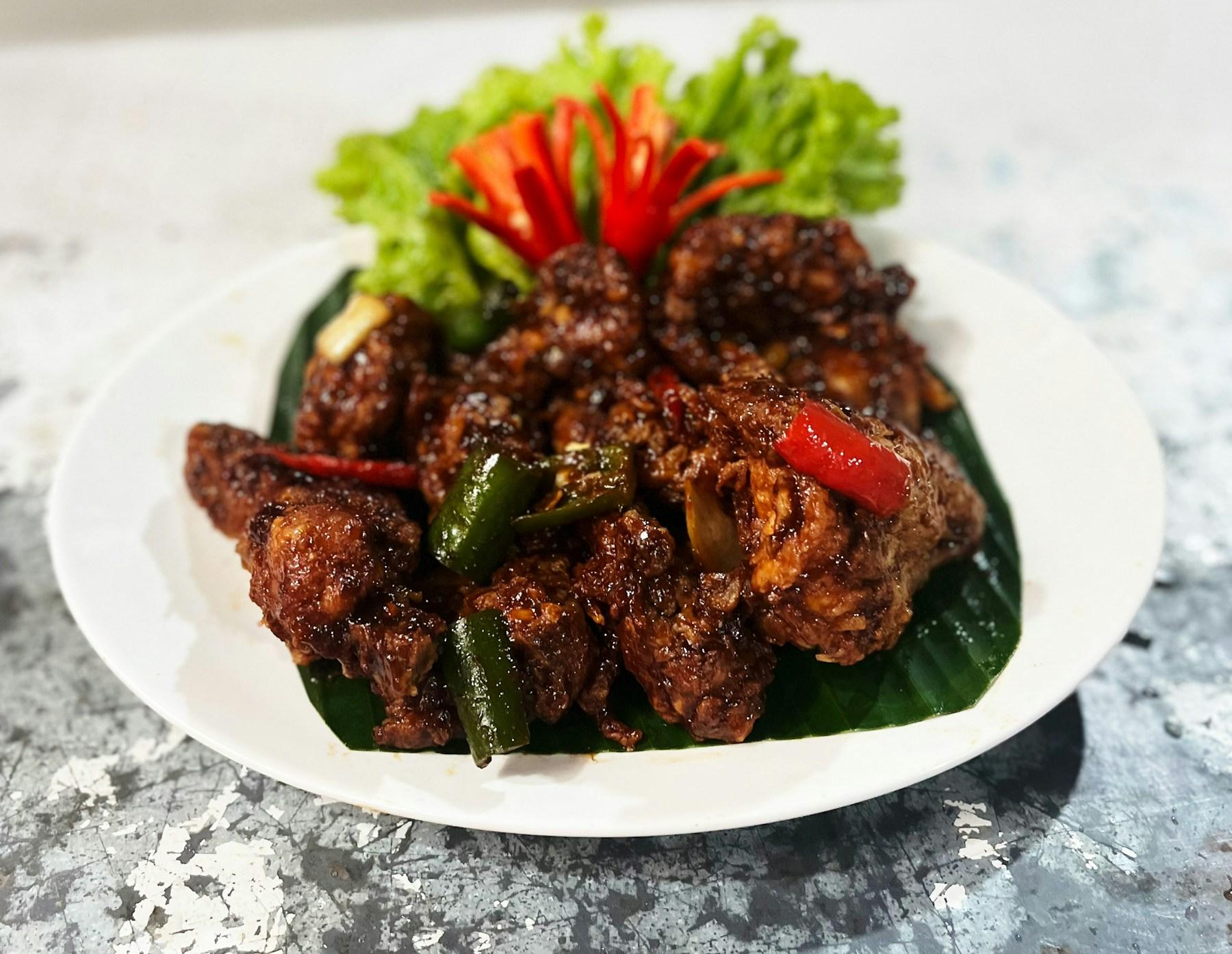 Dark, glossy spice-fried chicken piled on a banana leaf on a white plate, tossed with green and red chilli and finished with curled red chilli and frilled lettuce.