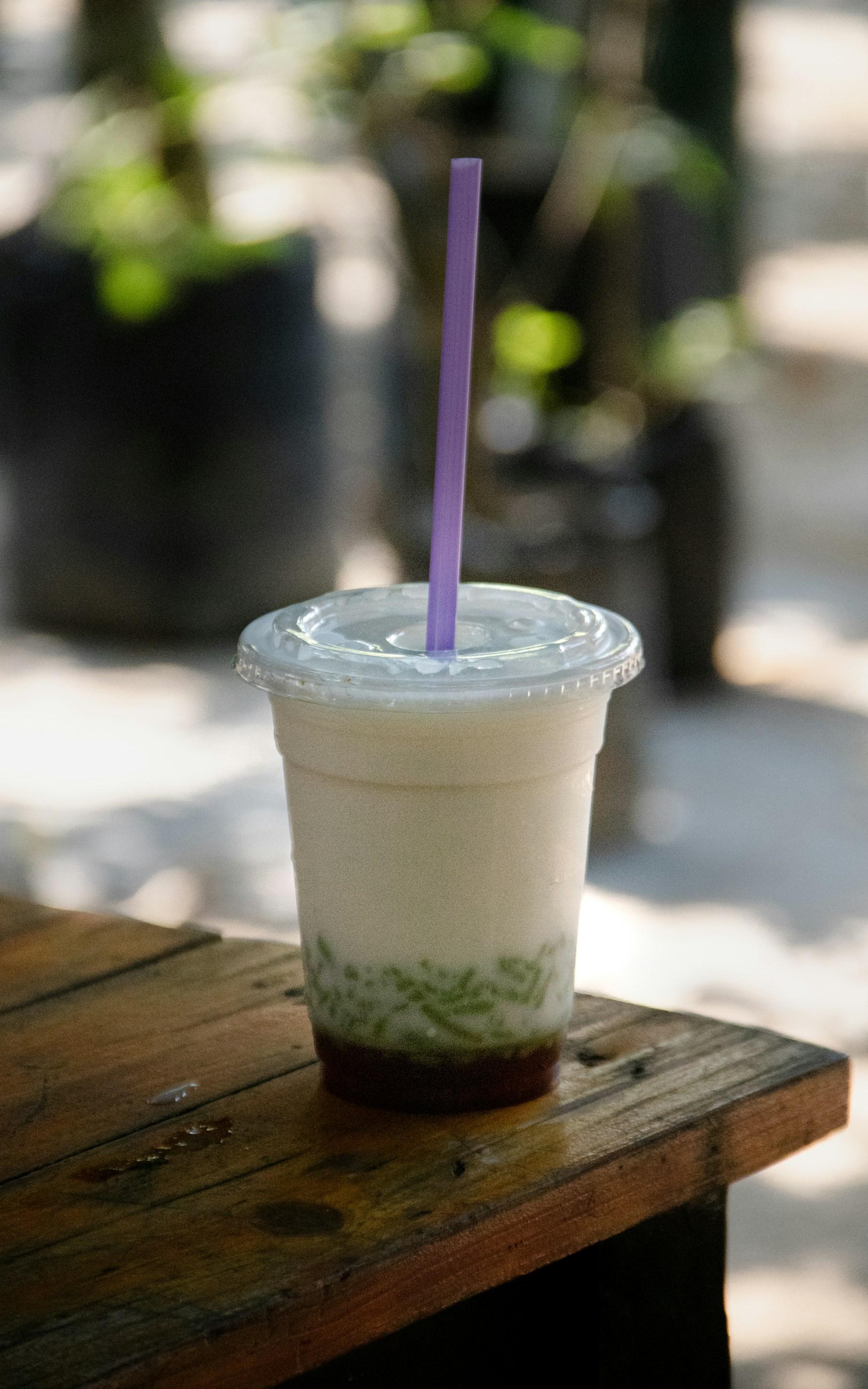 A takeaway cup of cendol on a sunlit wooden bench - green pandan jelly strands and dark gula Melaka settled beneath a tall pour of coconut milk, straw through the lid.