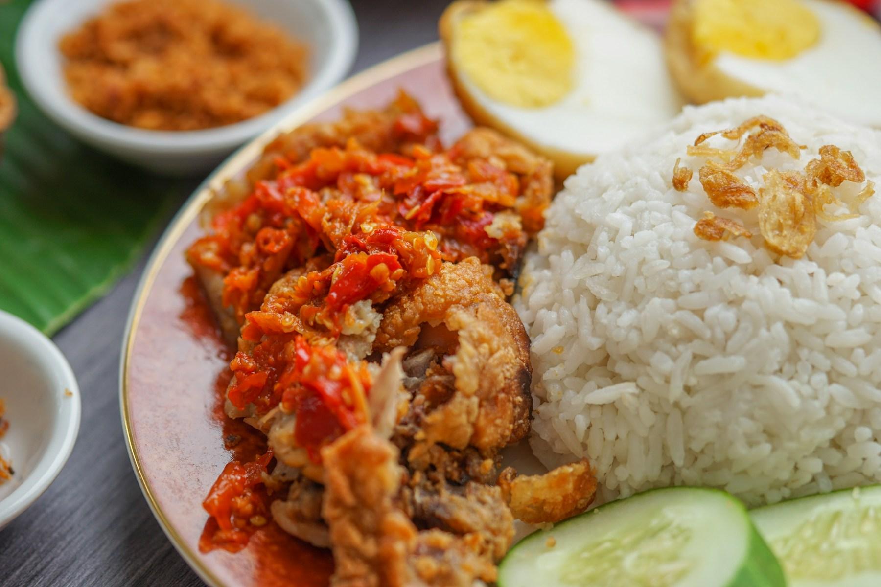 A plate of coconut rice scattered with fried shallots, served with fried chicken under a heap of pounded red chilli sambal, halved boiled eggs, cucumber slices and a side bowl of sambal.