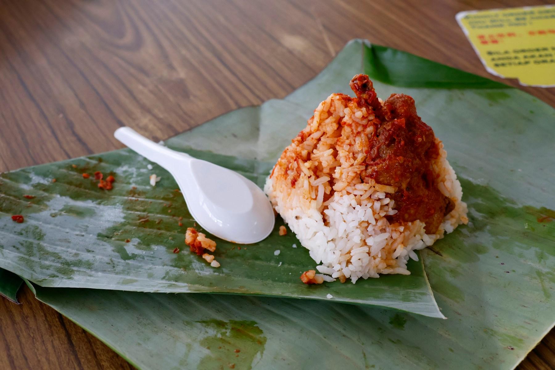 A mound of coconut rice opened out on a banana leaf, topped with dark red sambal tumis and a piece of meat in gravy, a white spoon set alongside.