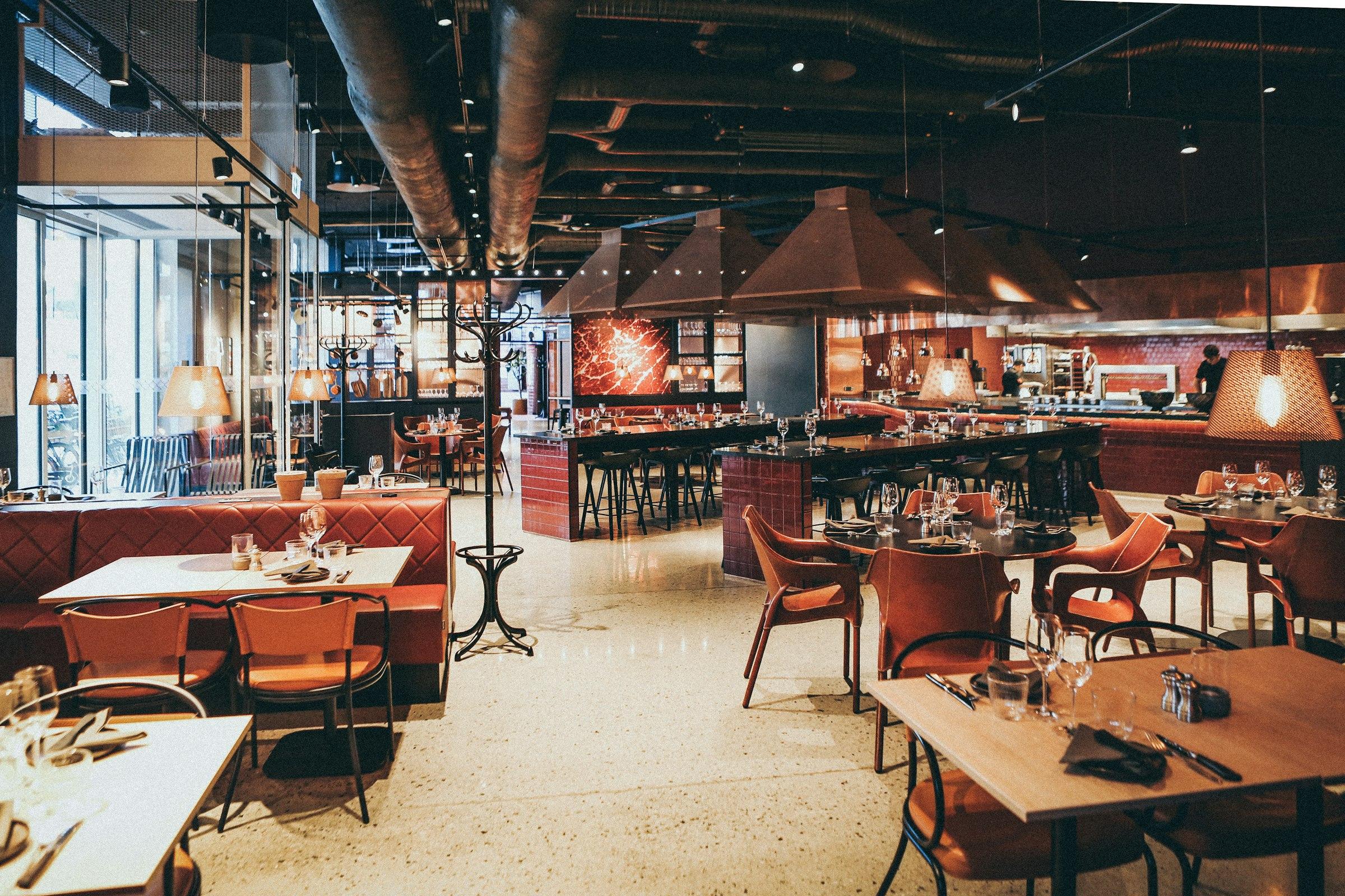 The warmly lit interior of a restaurant, with wooden tables, upholstered chairs and low pendant lamps.